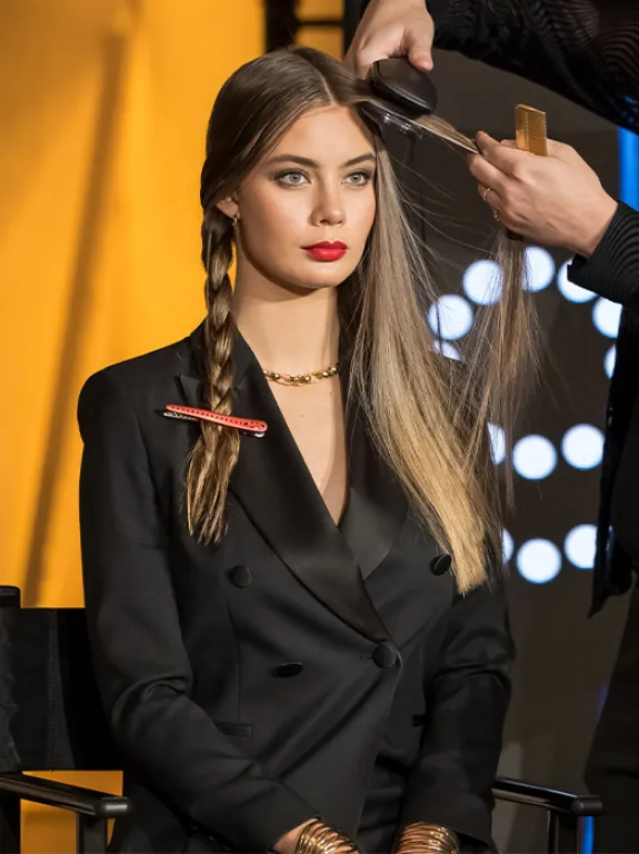 The model is sitting upright in a chair, one man is styling her hair, the other is filming the process with a camera