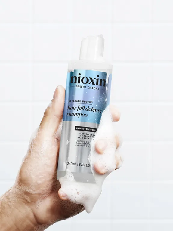 Male hand holding Hair Fall Defense Shampoo surrounded by foam, bathroom tiles in the background