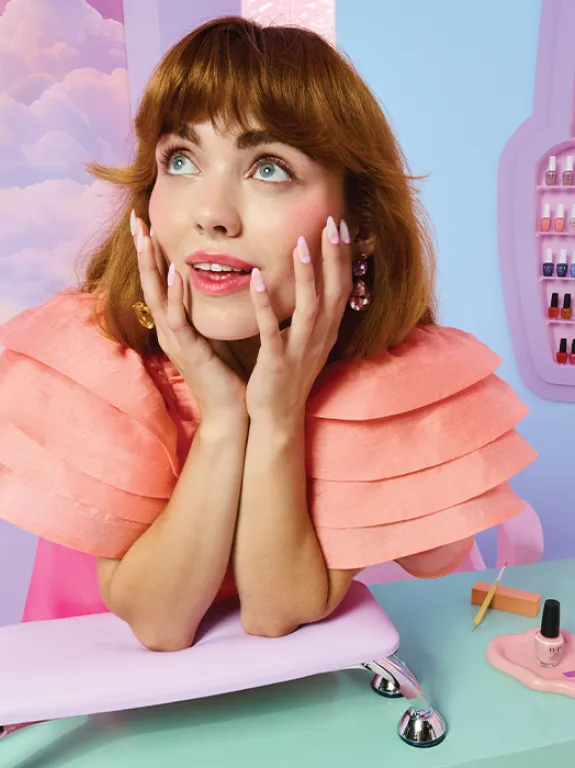 A model leaning on a manicure table in a pastel-colored room, with an OPI nail polish display in the background