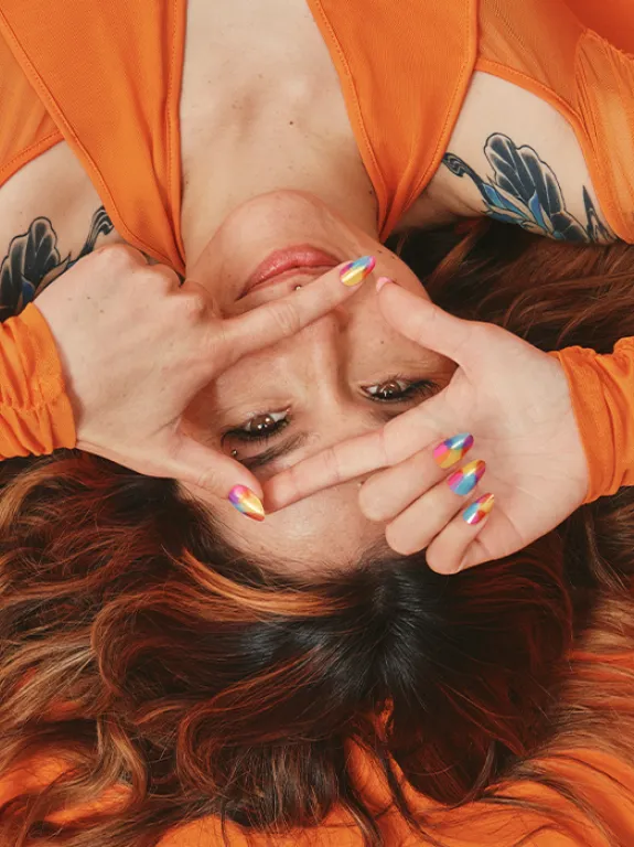 A brunette in an orange outfit lies on an orange floor, with her flowing, wavy hair spread out, making a gesture as if she is holding a camera