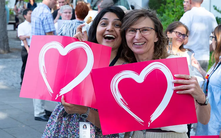 WellaCompany-Card-01-Join-Us.jpg Two smiling women hold red boards with a heart logo, crowd of people in the background