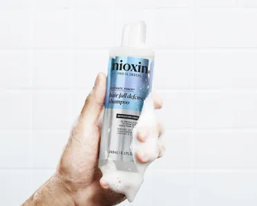 Male hand holding Hair Fall Defense Shampoo surrounded by foam, bathroom tiles in the background