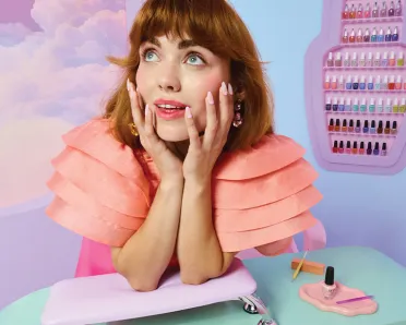 A model leaning on a manicure table in a pastel-colored room, with an OPI nail polish display in the background