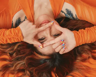 A brunette in an orange outfit lies on an orange floor, with her flowing, wavy hair spread out, making a gesture as if she is holding a camera
