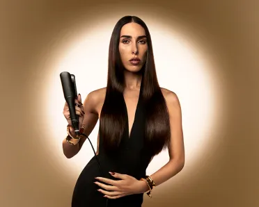 Brunette holding ghd hair styler; golden brown background with circle of light in the center